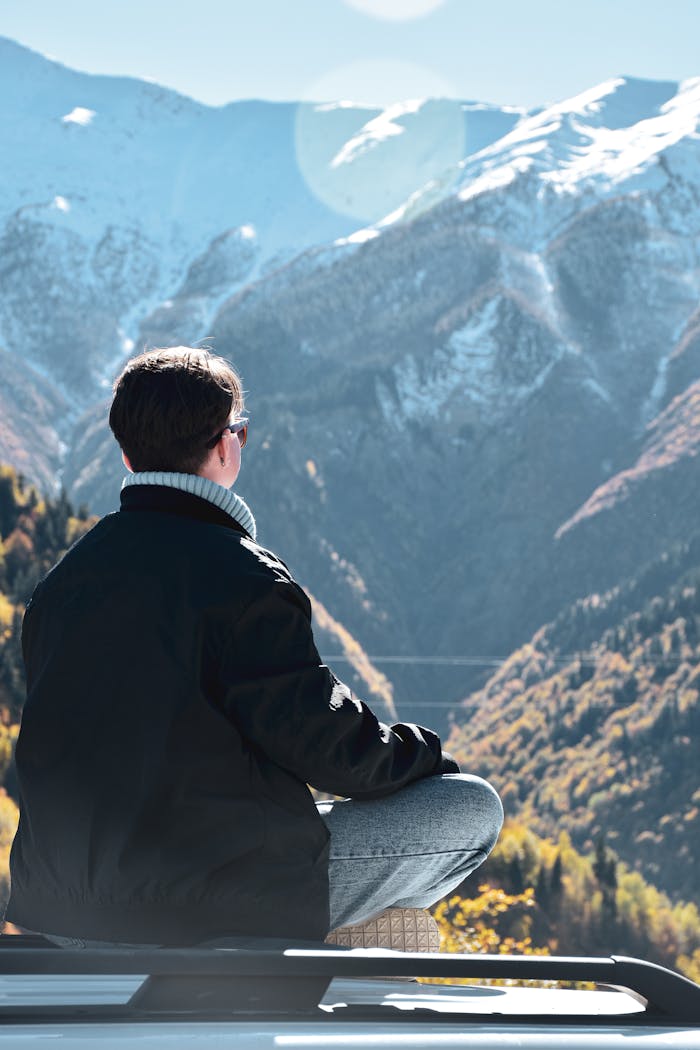 A woman enjoys solitude and scenic mountain views, sitting on a car roof under the sunshine.