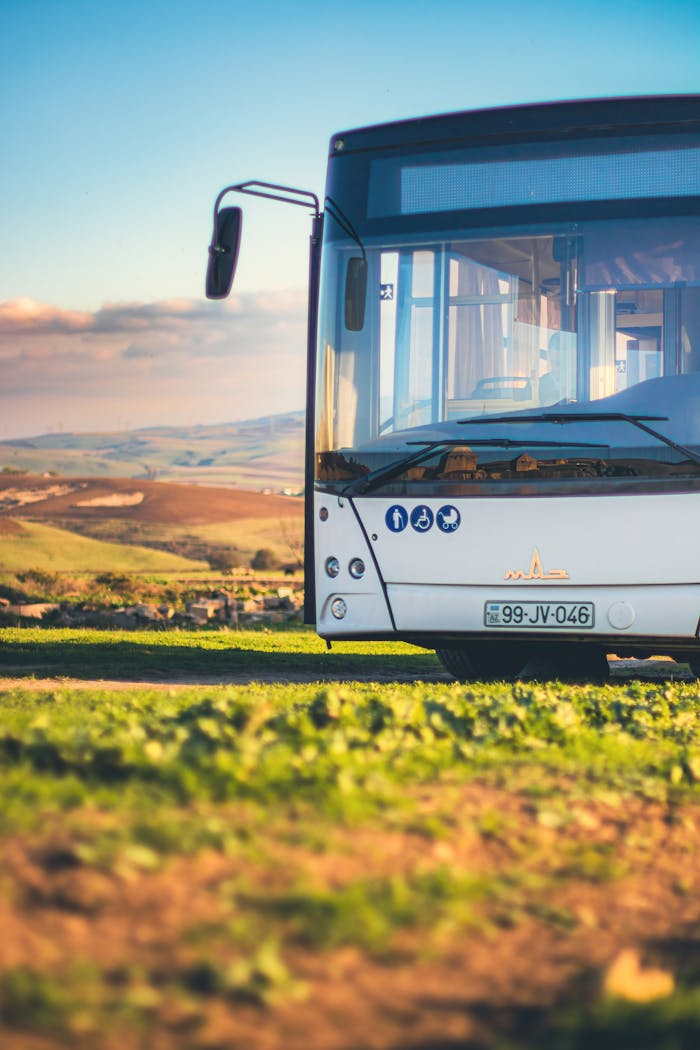 A white bus parked in a sunny field with rolling hills and a clear sky, offering a scenic countryside view.